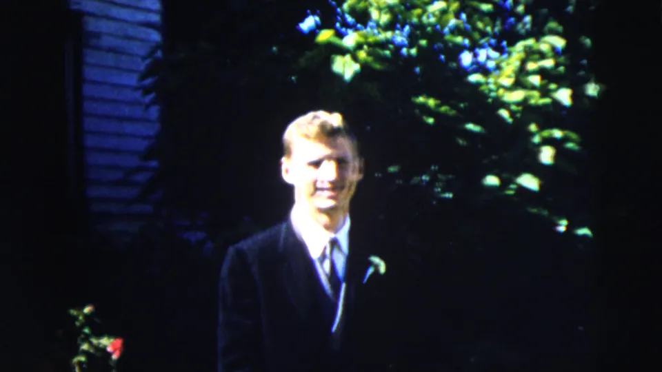 A wider 1962 Moline home-movie frame shows the young man in formal clothes standing outside near siding and garden foliage.