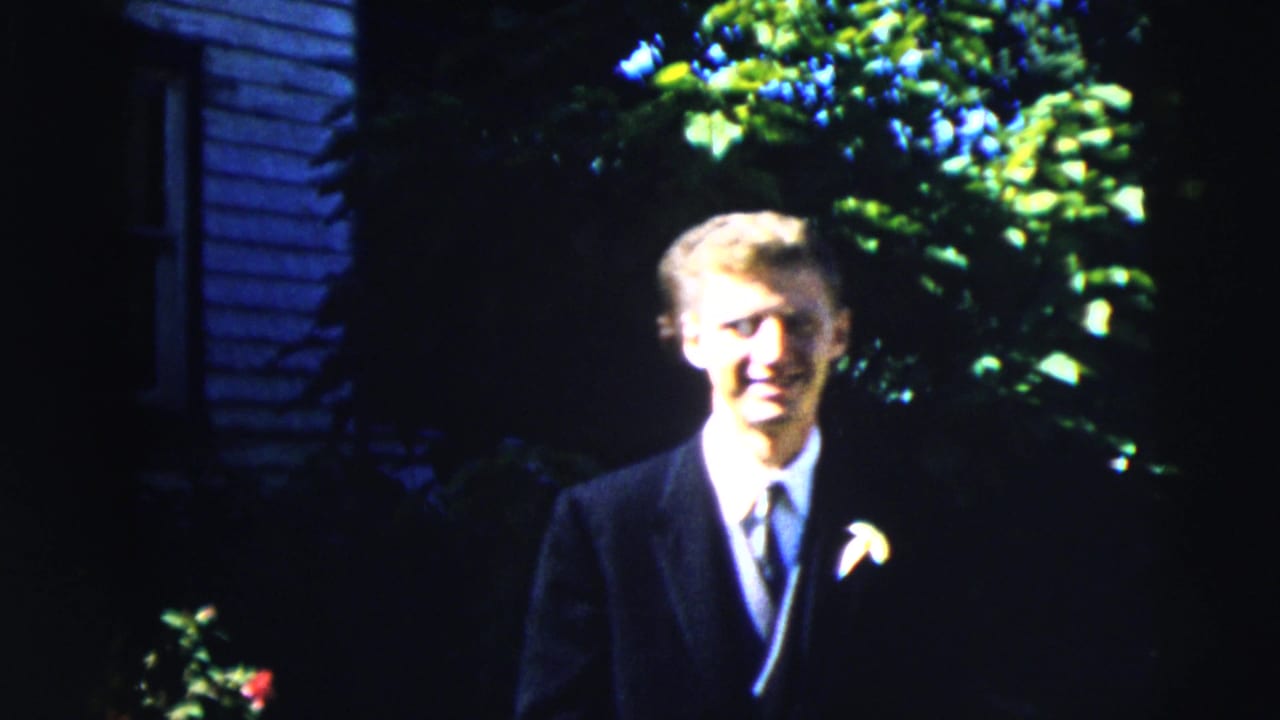 In metadata-supported 1962 Moline footage, a young man in a dark suit and boutonniere stands outside before greenery and blue siding.