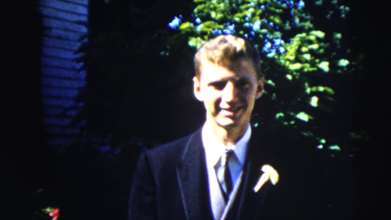 The suited man smiles toward the camera, moving closer in the 1962 Moline home-movie shot with foliage and siding behind him.