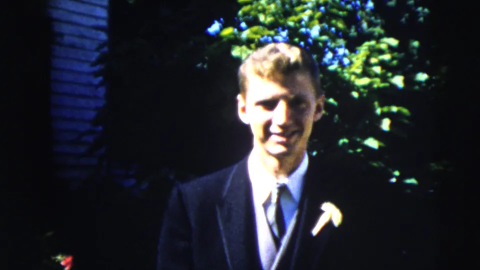 The suited man smiles toward the camera, moving closer in the 1962 Moline home-movie shot with foliage and siding behind him.