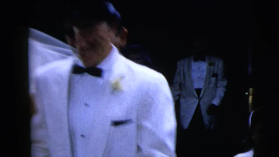 A handheld close view catches the groom in a white dinner jacket and bow tie as figures remain in the dark background.