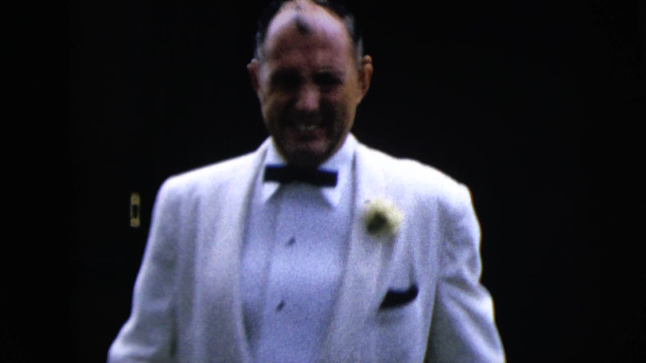 Groom in a white tuxedo walks toward the camera in a dark interior.