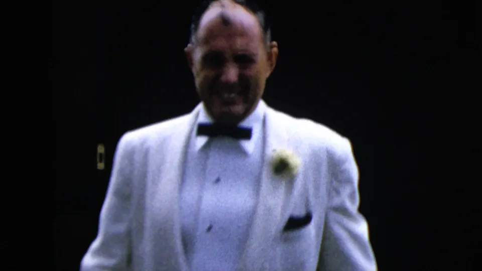 In 1962 Moline wedding footage, a groom in a white tuxedo and black bow tie walks forward as a bridesmaid stands behind him.