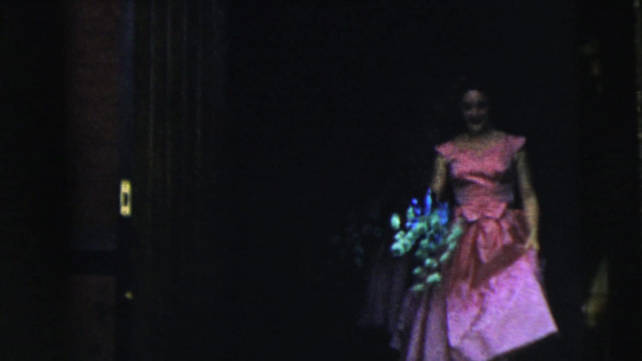 Bridesmaid in a pink dress carries a bouquet inside a dark doorway.