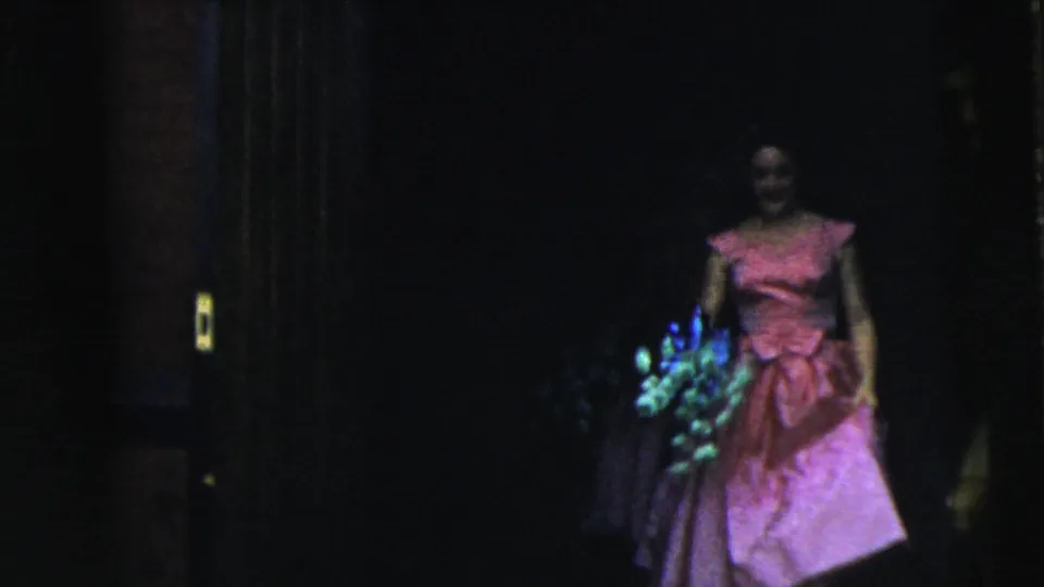 A bridesmaid in a pink dress and purple hat carries blue-green flowers in the 1962 Moline wedding home movie.