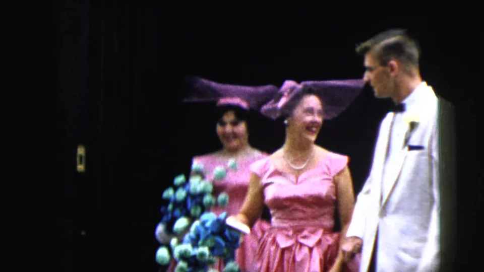 Two pink-dressed bridesmaids smile and move forward beside a man in a white tuxedo during the 1962 Moline wedding procession.