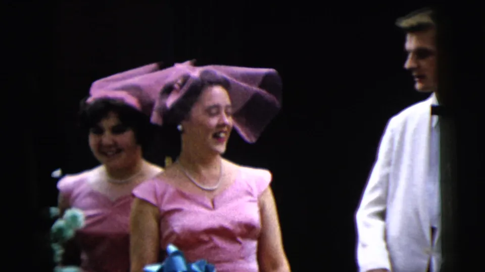 A smiling woman in a pink dress and large purple hat passes another attendant and a white-tuxedoed man in the 1962 wedding footage.