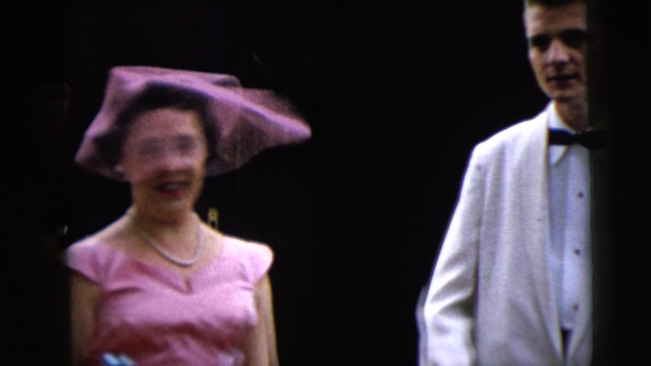 1962 Moline home-movie frame showing a woman in pink formalwear and hat near men in white dinner jackets at a dark entrance.