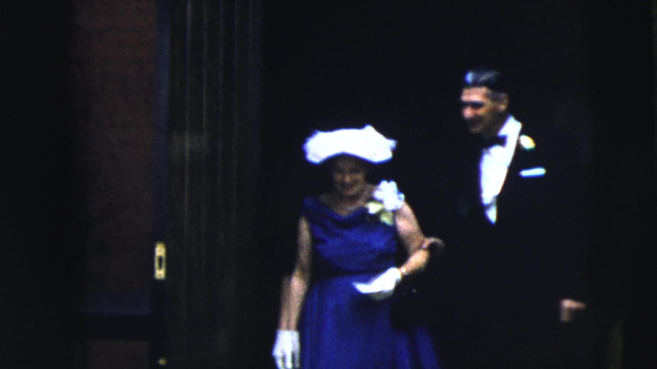 Woman in a blue dress and white hat walks from a dark doorway with a man in a tuxedo.