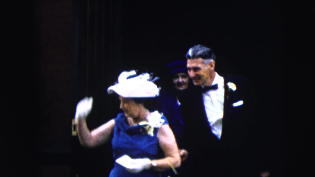 The woman in a blue dress and white hat lifts a gloved hand near two men in formal attire in the Moline 1962 home-movie scene.
