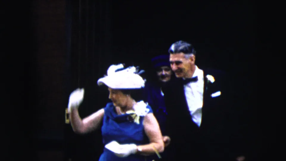 The woman in a blue dress and white hat lifts a gloved hand near two men in formal attire in the Moline 1962 home-movie scene.