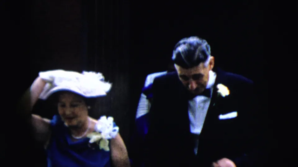 The woman holds onto her white hat as she walks beside a tuxedoed man, with dim doorway shadows behind them in the 1962 Moline footage.