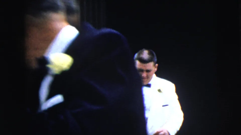 A blurred formalwear figure crosses the foreground while a man in a white dinner jacket stands farther back in the 1962 Moline home movie.