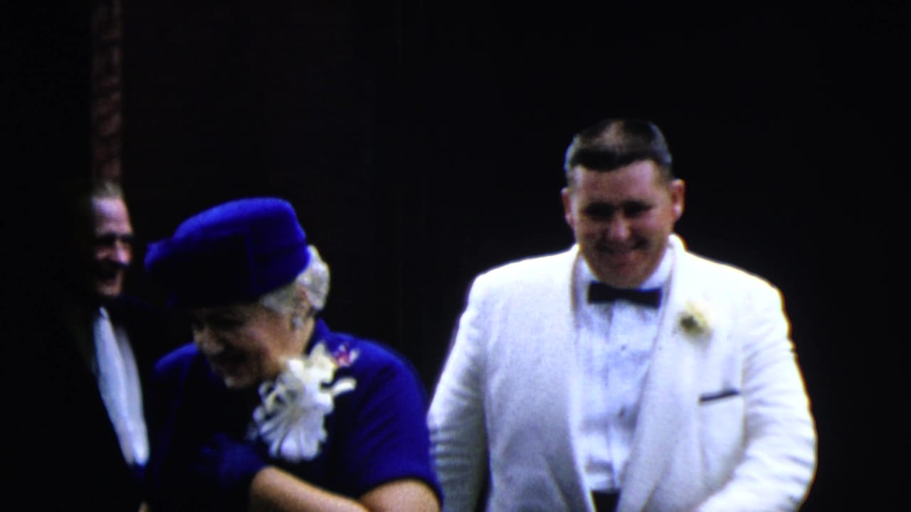 Adults in formal clothes near a dark entrance, with a man in a white tuxedo facing the camera.