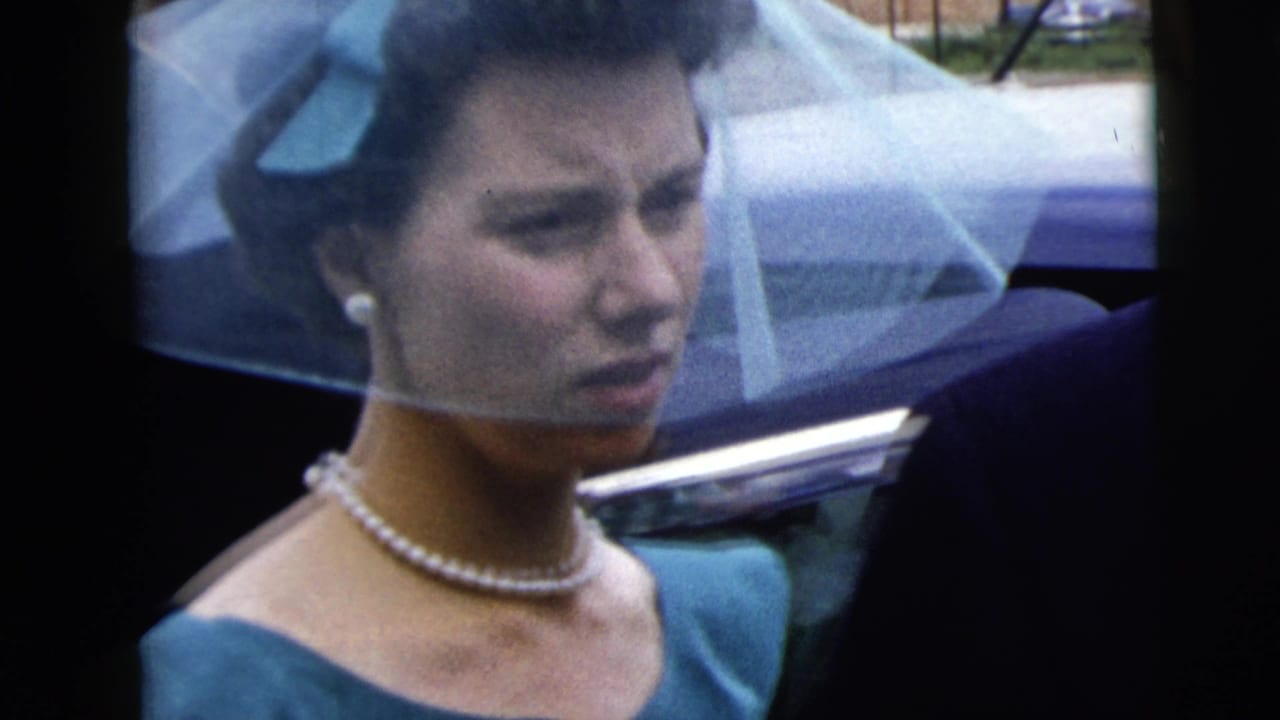 Veiled woman in teal dress and pearls beside a parked car.