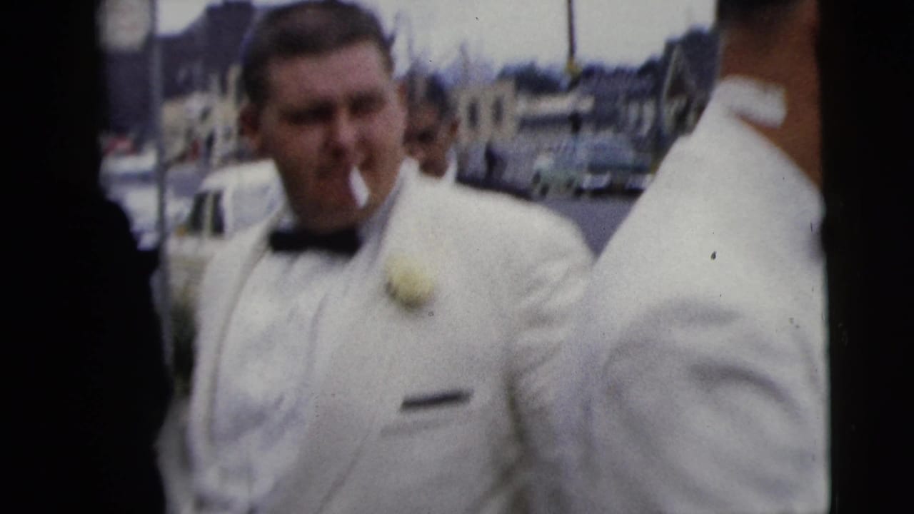 Man in white tuxedo with boutonniere stands near parked cars.