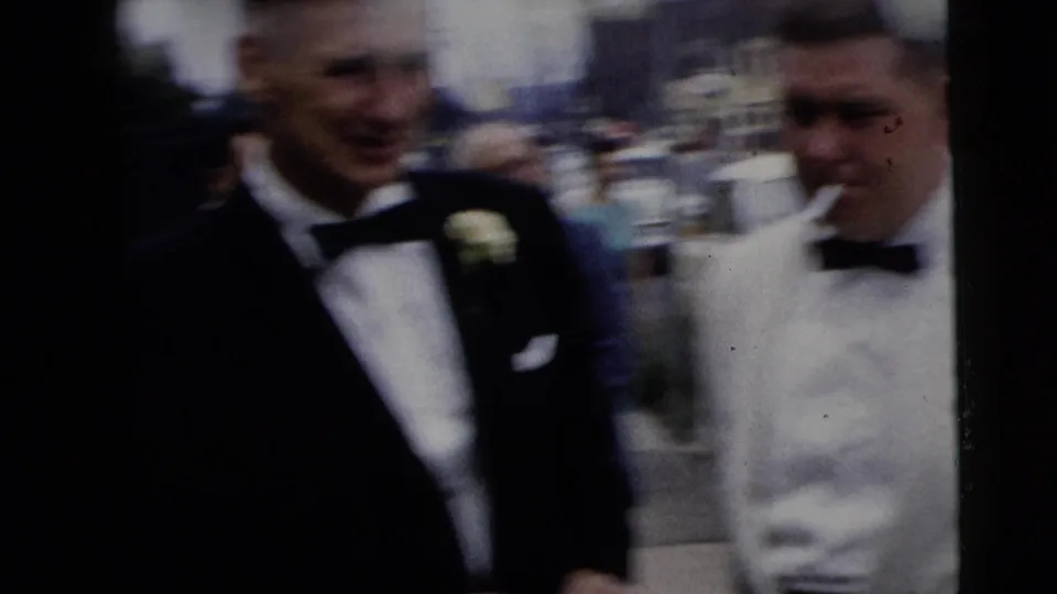 Two men in wedding formalwear, one in a dark tuxedo and one in a white tuxedo, move through a parking lot in the 1962 home movie.