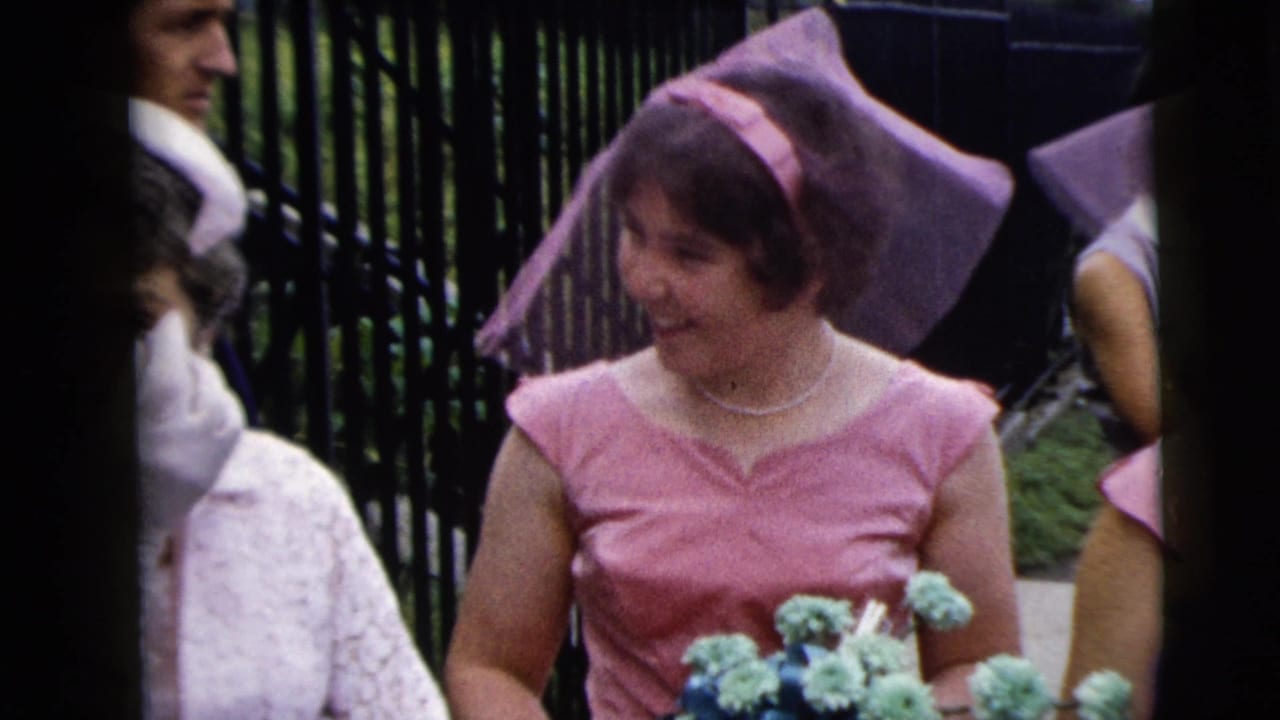 Two bridesmaids in pink dresses and veils walk past guests beside an iron fence.