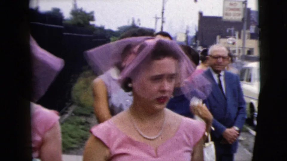 A pink-dressed bridesmaid with a matching veil and pearl necklace moves through an outdoor wedding crowd in 1962 Moline footage.