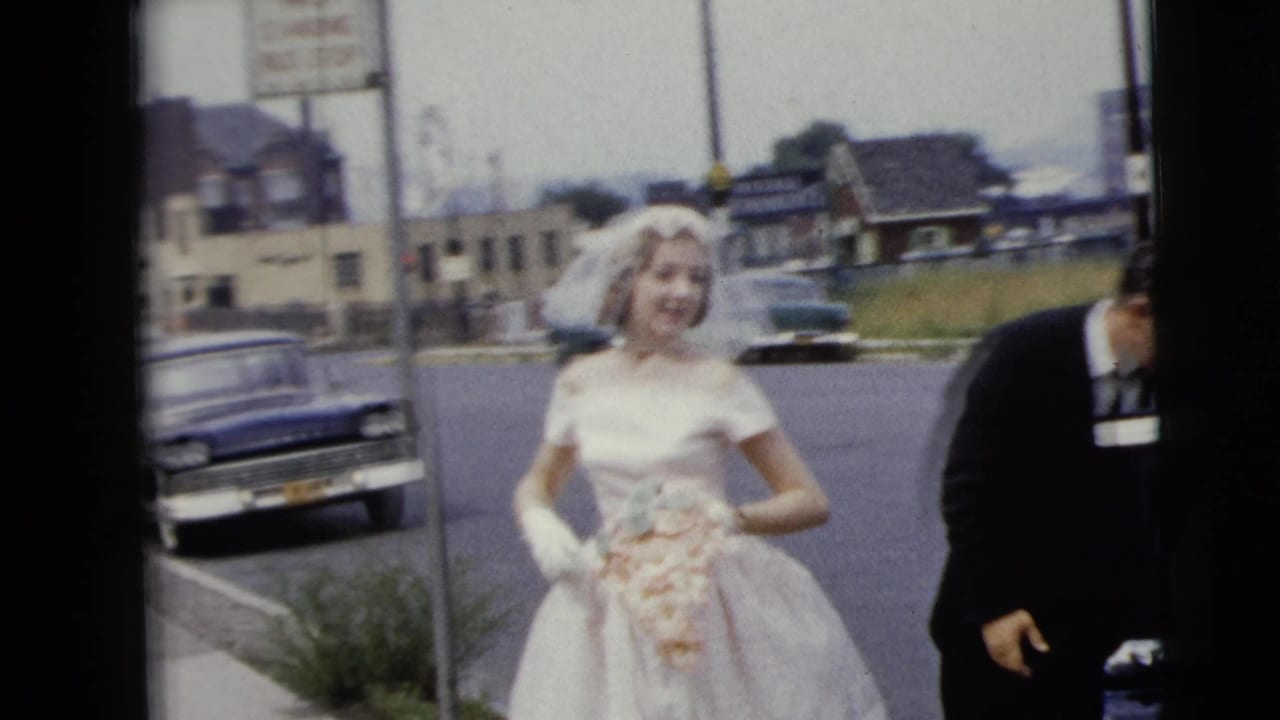 Bride in a white gown holds flowers near a street as a man in a dark suit stands beside her.