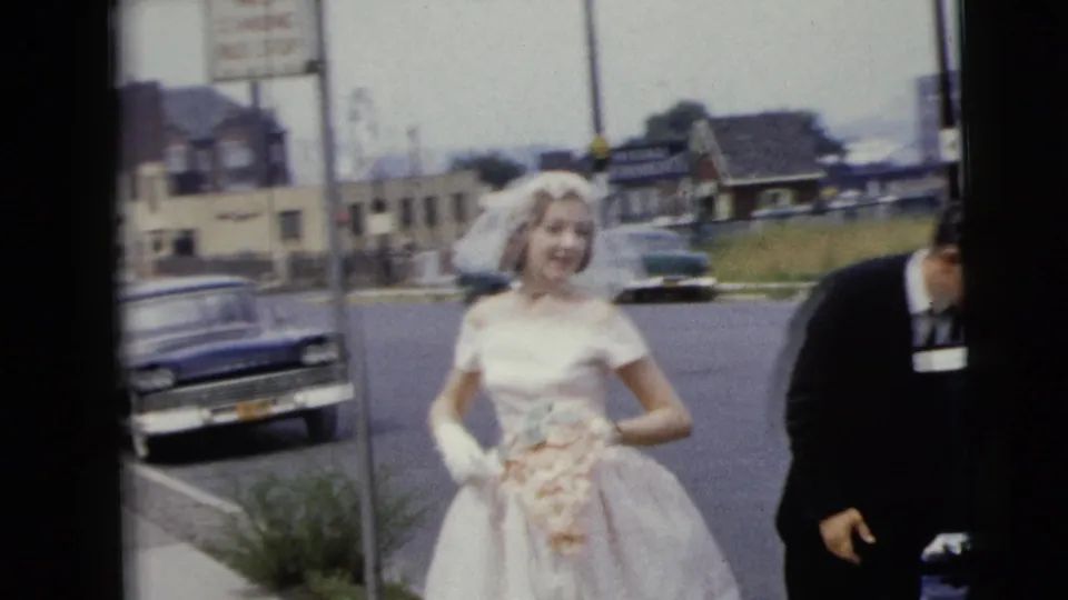 In Moline, Illinois, 1962 home-movie footage, a bride in a white gown and veil holds a bouquet near parked cars while a man assists beside her.