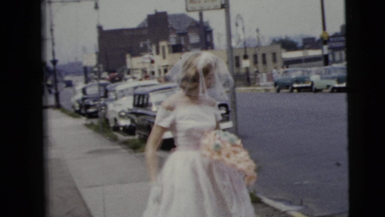 A bride in a white gown and veil walks on a Moline street in 1962, holding a pastel bouquet with parked cars and buildings behind her.