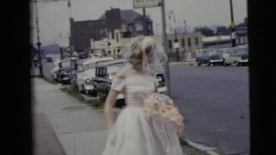 A bride in a white gown and veil walks on a Moline street in 1962, holding a pastel bouquet with parked cars and buildings behind her.