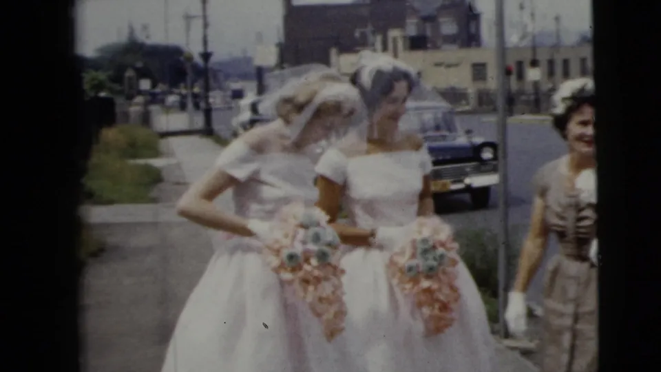 Two brides in white off-the-shoulder gowns and veils stand close together with bouquets on a Moline street, joined by a woman in formal dress.