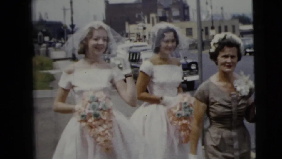 Two brides in white gowns hold bouquets beside an older woman in a brown dress on a Moline street, with early-1960s cars in the background.