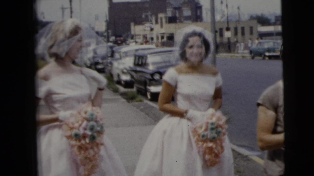 Two women in white dresses and veils hold pastel bouquets on a city sidewalk.