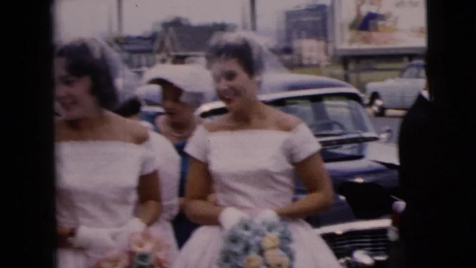 Two bridal party members in white dresses and veils walk side by side past parked cars on a 1962 Moline city street.