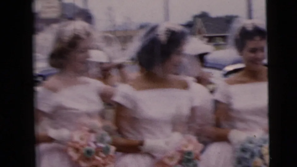 A softly blurred home-movie frame shows bridal party members in veils and white dresses moving together on a Moline street.