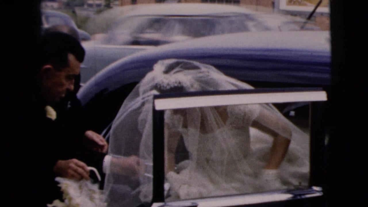 Bride in a veil leans from an open blue car while a man in a dark suit stands nearby.