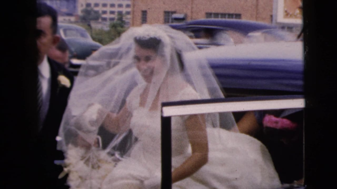 The veiled bride is upright beside the open car door in the 1962 Moline wedding scene, holding flowers as cars and brick buildings sit behind her.