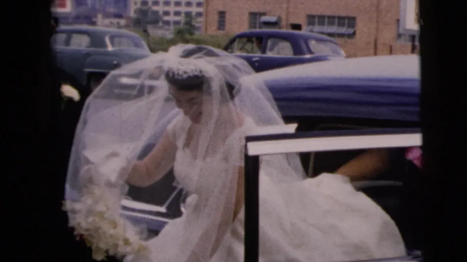 The bride in veil and white dress lowers her gaze near the open car door, with attendants and parked mid-century cars visible in Moline, Illinois.