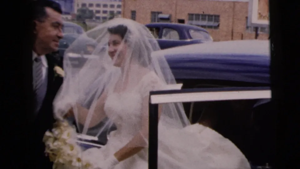 The bride smiles through her veil beside a suited man and the open blue car door during the 1962 wedding arrival in Moline, Illinois.