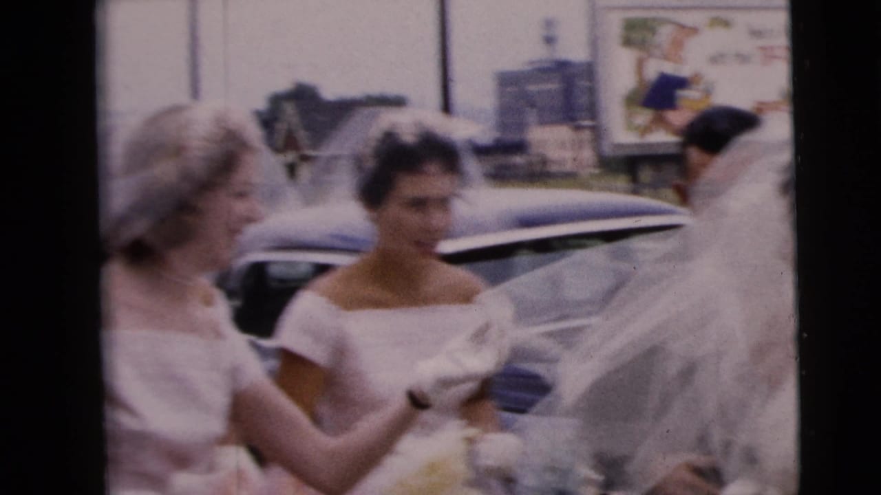 Women in formal dresses stand near the veiled bride beside parked cars.