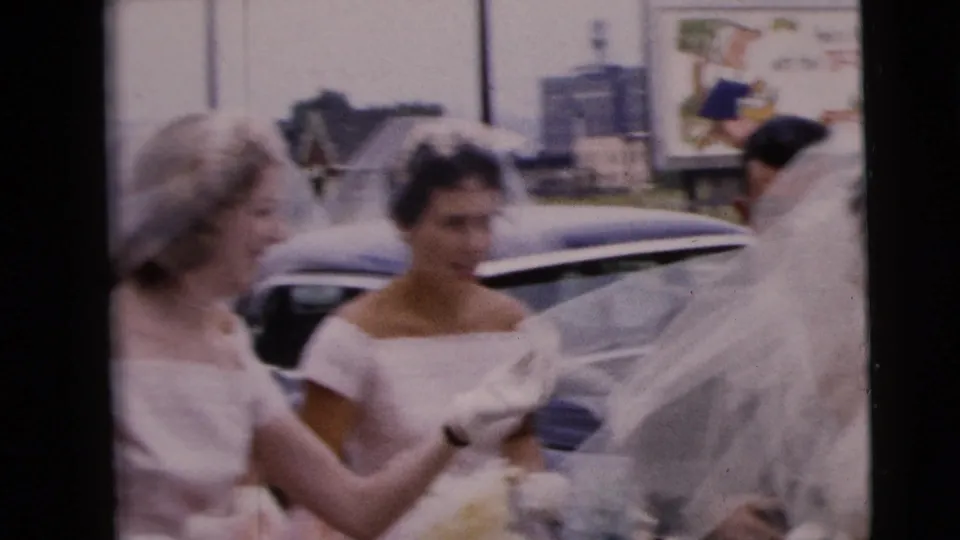 Women in wedding attire gather around the veiled bride on a Moline street with 1960s cars and signage behind them.