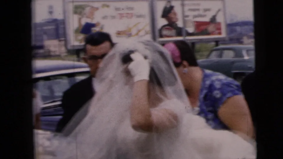 The bride raises a hand to her veil as helpers assist beside the wedding car in the 1962 Moline street scene.