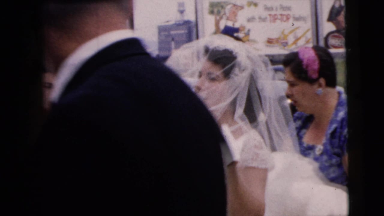 Bride in veil beside a woman in blue floral dress as a suited man passes in the foreground.