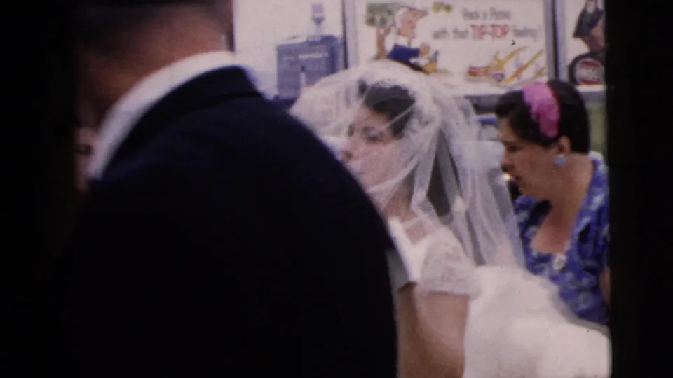 In Moline, Illinois, 1962, a veiled bride stands on a sidewalk while a woman in a blue floral dress tends to her gown and a suited man crosses close to camera.