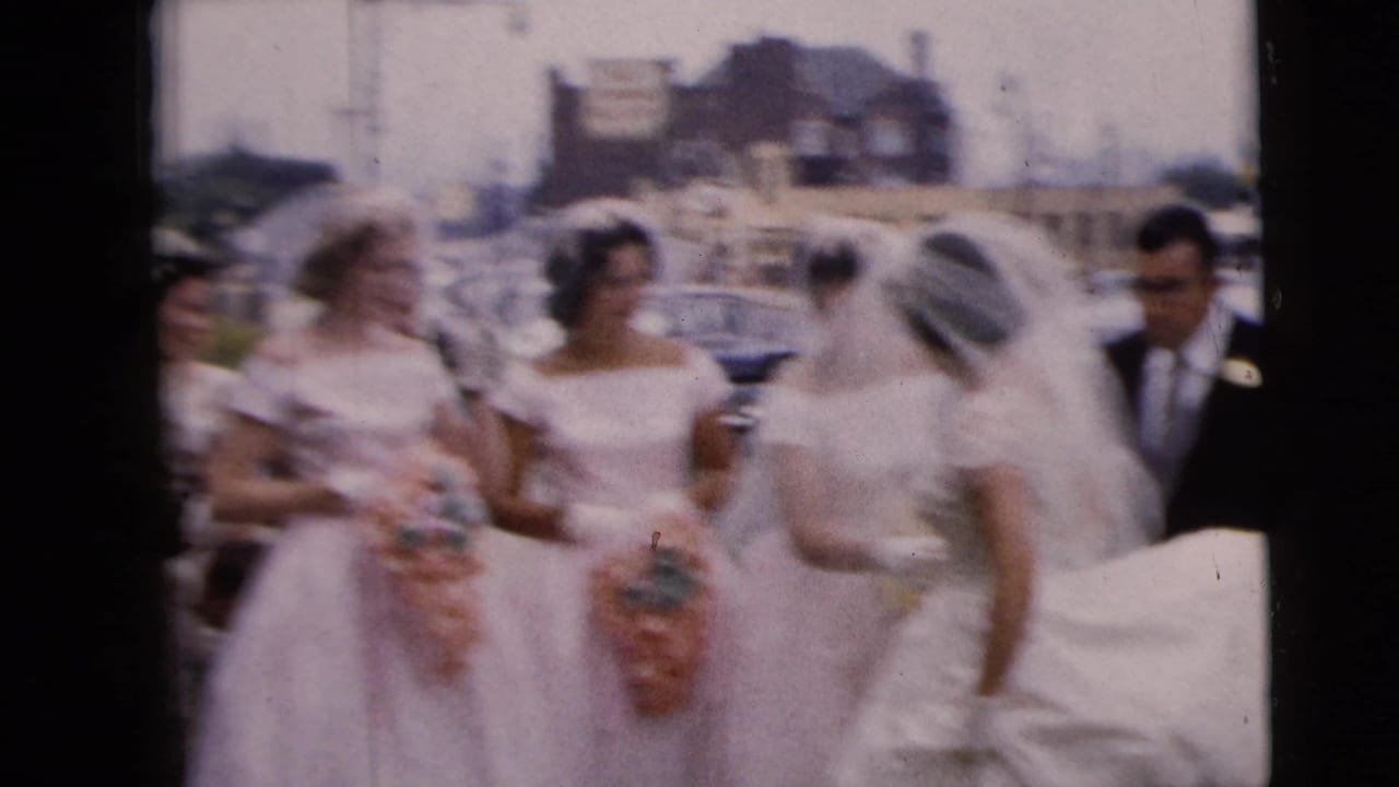 Bride and bridesmaids in pale dresses stand near parked cars on a commercial street.