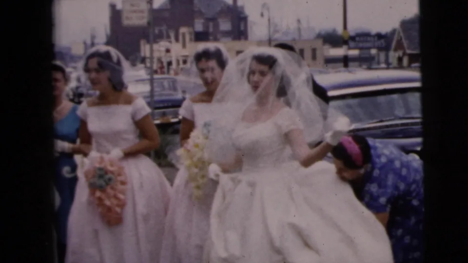 On a Moline sidewalk in 1962, a woman bends to adjust the bride’s white train as bridesmaids with bouquets wait beside parked cars.