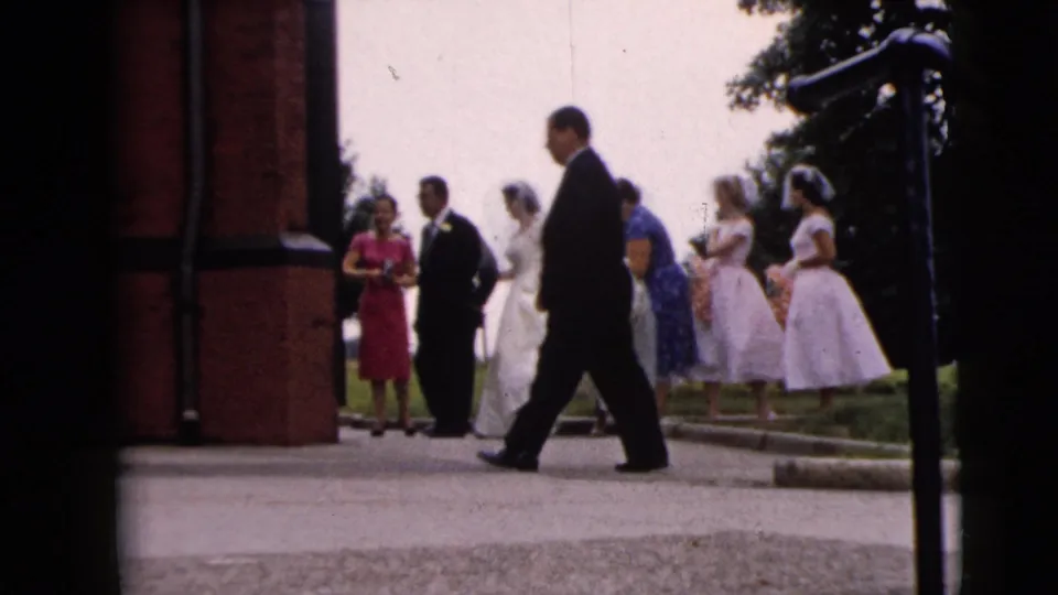 A wedding group stands near the brick church entrance as a man in a dark suit walks across the foreground in 1962 Moline footage.