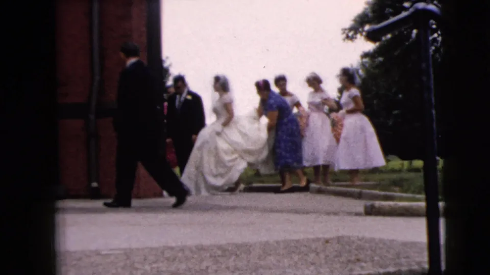 The bride, groom, and bridesmaids remain outside the brick church as a suited man crosses the frame in the Moline wedding home movie.