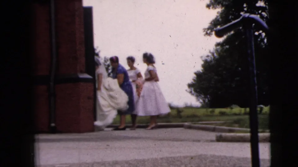 A 1962 home-movie frame shows attendants helping the bride's gown as the group moves beside the church entrance.