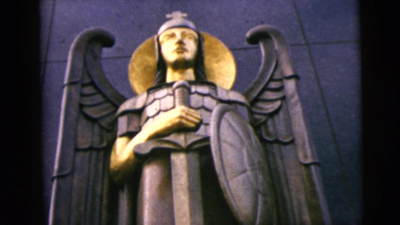 Armored angel statue with gold halo, wings, sword, and shield against a dark wall.