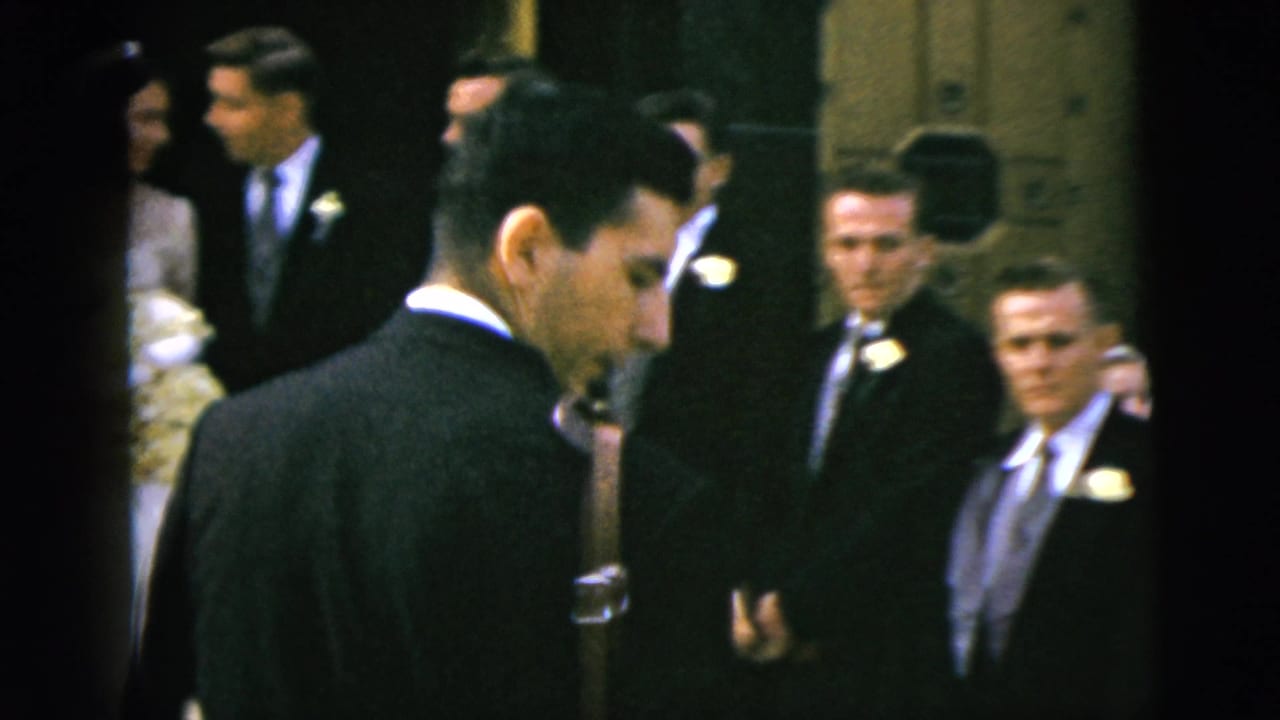 Men in dark suits stand near a wedding party partly obscured by foreground figures.