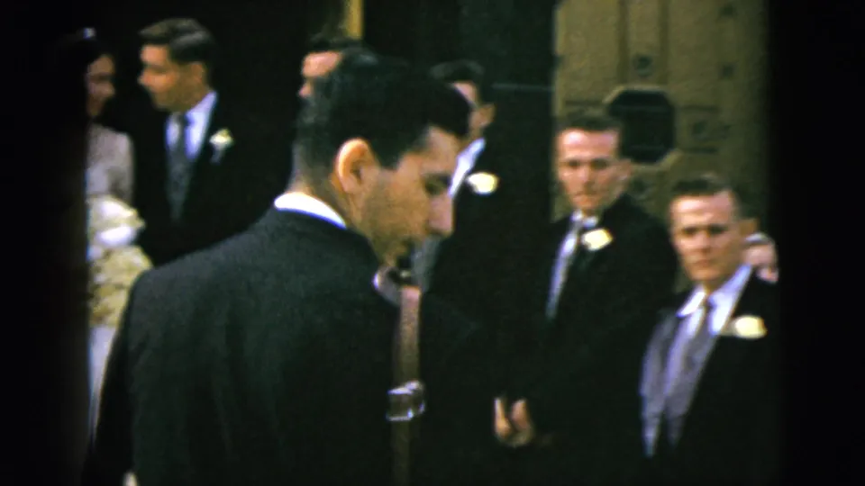 Wedding party in Moline appears partly blocked by men in dark suits as the group waits indoors for pictures.