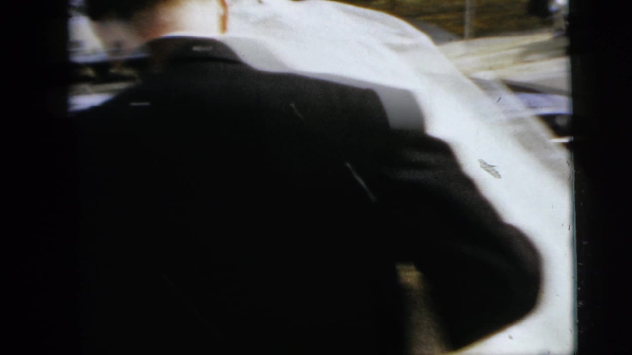 Bride and groom seen from behind near a car with a white-gloved arm crossing the frame.